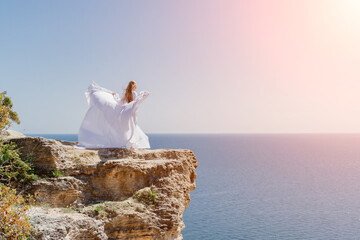 Fototapeta na wymiar Woman in a white dress on the sea. Side view Young beautiful sensual woman in white long dress posing on a rock high above the sea at sunset. Girl in nature against the blue sky