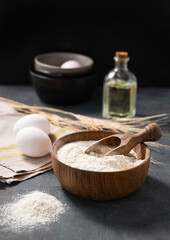 Wheat flour in a wooden bowl with a scoop on a dark blue background with eggs, oil and dry branches wheat. Organic ingredients for baking.
