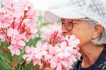 Closeup on senior woman in white cap and eyeglasses smelling pink oleander flower