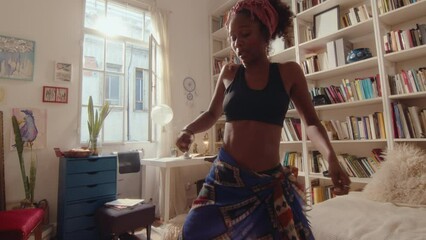 Young carefree African-American girl dancing and enjoying the music in a sunlit living room, having fun during the day at home. Zoom shot, full length
