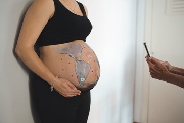 Tender scene of a multi-ethnic lesbian couple photographing the pregnant woman's belly drawing at...
