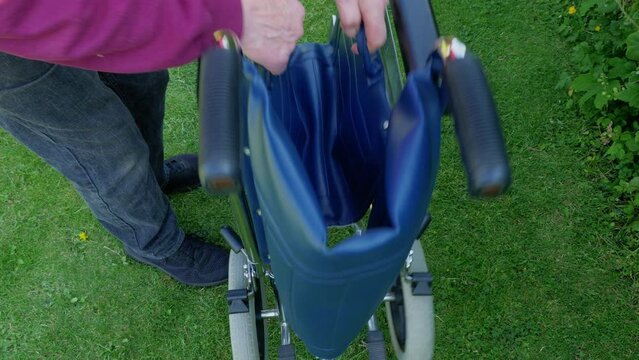 Closeup of a man&rsquo;s hands grabbing two handles on the fabric seat of an empty, portable wheelchair, outside on grass, causing it to fold into a much narrower size, ready to be moved.