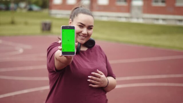 Portrait of smiling overweight girl on basketball court showing green screen phone to camera