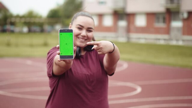 Smiling overweight young woman  showing green screen mobile phone by pointing finger by looking camera outdoors after training or sport