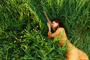 a relaxed woman enjoys summer lying in the tall green grass in a long orange dress stretched out...