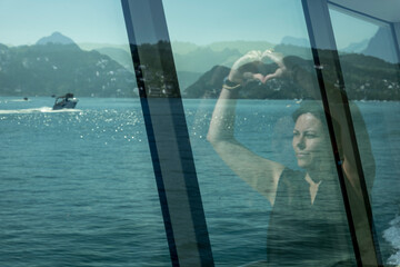 Woman Reflected in the Window on a Passenger Ship and Making a Heart Shape in a Sunny Summer Day on Lake Lucerne with Mountain in Switzerland.
