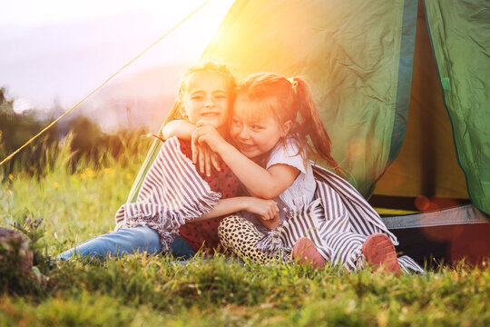 Two little hugging girls sisters sitting on the green grass next camp tent entrance, cheerfully smiling. Careless childhood, family values and outdoor activities concept photo.