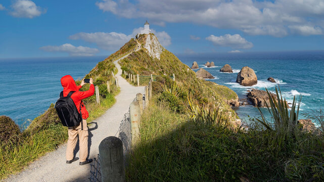 Young tourist man in red hood using smartphone take picture at Nugget Point Lighthouse, South Island, New Zealand