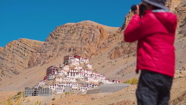 Defocused Indian male tourist taking picture with camera in front of the Key monastery in front of the Himalayan mountains near Kaza in Spiti Valley, Himachal Pradesh, India. Tourist taking photo. 