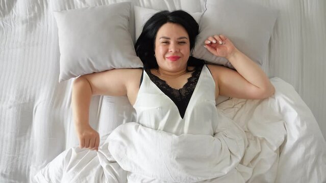 Woman with dark long loose hair wakes up pleasantly in cozy bedroom. Plump housewife lies on white double bed ready for starting new day, top view