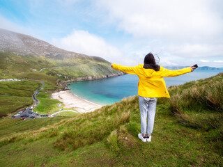 Teenager girl in yellow jacket standing on a hill and looking at stunning nature scenery of Keem...