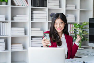 Asian business woman celebrating victory success Excited Success. Woman using laptop and smartphone at work inside office holding hand up and happy triumph gesture.