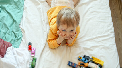 Fototapeta premium Top view on cute baby boy lying in bed with lots of toys and cars. Child development, education and entertainment