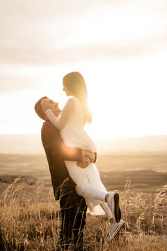 Pareja joven de novios recien casados celebrando su boda informal con una sesion de fotos al atardecer en el campo, el la coge a ella y se miran con amor