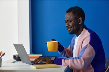 Vibrant side view portrait of black man using laptop at workplace in office with clock on blue wall, copy space