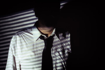Fototapeta na wymiar Faceless Shadow Man Interrogator. Mysterious, anonymous man with face hidden in shadows of blinds, wearing white shirt and black tie. Shot in film noir style.