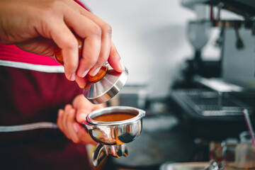 Hand of a barista holding a portafilter and a coffee tamper making an espresso coffee. Barista presses ground coffee using a tamper in a coffee shop