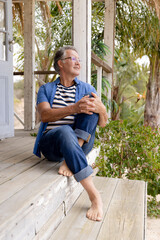 Smiling caucasian senior man dressed in casuals contemplating while sitting on steps outside cottage