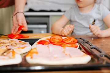 Close-up of mother and daughter cooking food together in the kitchen, selective focus on mini pizzas with tomatoes