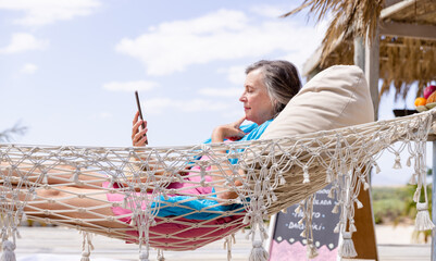 Side view of caucasian senior woman using digital tablet and lying on hammock at beach against sky