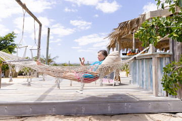 Side view of caucasian senior woman using digital tablet and resting on hammock at beach against sky