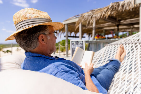 Caucasian senior man wearing hat and reading book while lying on hammock at beach
