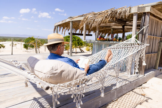 Caucasian senior man wearing hat and reading book while lying on hammock at beach against sky