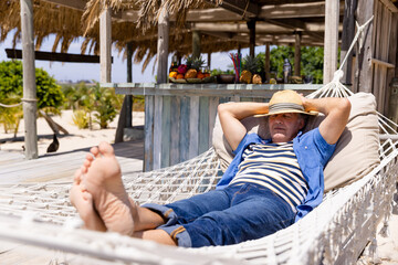 Caucasian senior man with hands behind head sleeping on hammock at beach