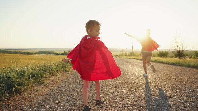 Carefree children play superheroes wear red cloaks run along rural road on nature with spreading flying hands.Teen girl sister kid boy brother have game together. Happy childhood, siblings friendship.