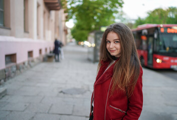 Beautiful Young Girl is Standing on the side Walk in Vilnius Old Town, Lithuania. Wearing Red jacket and Black Trousers. Beautiful Spring Day. Smiling. Public Transport Bus in Background