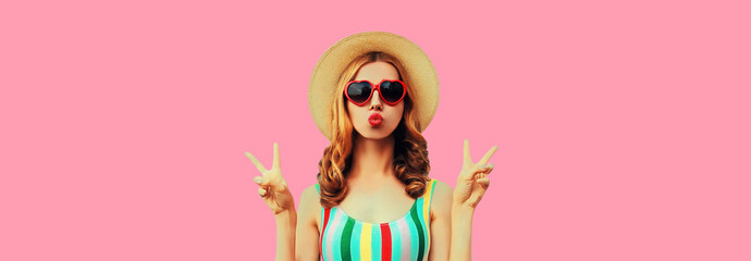 Portrait of beautiful young woman model blowing her lips and posing wearing summer straw hat, red heart shaped sunglasses on pink background