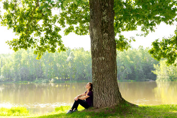 Young woman sitting on grass under high big oak,trunk tree near lake,river,meditating,relaxing in...