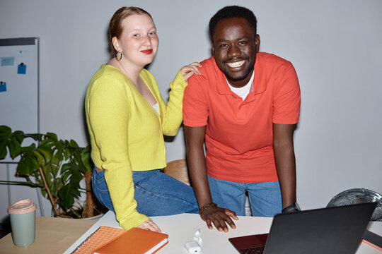 Front view portrait of smiling two people looking at camera at workplace and leaning on desk, shot with flash