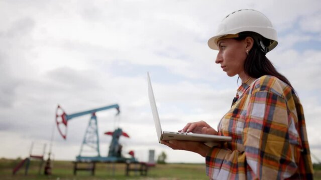 Oil industry. Engineer stand in field next to an oil rig station working with laptop computer and typing text to keyboard. Oil production business finance concept. Gas production. Worker in helmet.