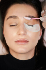 Woman with eye patch under lower eyelid keeping eyes closed while beautician applying cleansing mousse on eyebrows with lash brush. Female person having eyelash extension procedure in beauty salon.