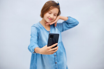 Naklejka premium Smiling little girl in casual denim dress hold smartphone do selfie