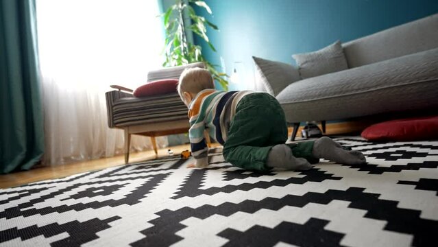 Back view of the unrecognizable boy in casual clothes, craws on floor and playing car toys while spending time in cozy living room at home