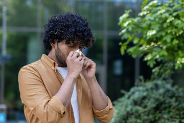A young hispanic man stands on the street and wipes his nose with a tissue. He has a runny nose, flu, suffers from seasonal allergies
