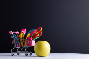 Close up of miniature shopping trolley school materials, apple and copy space on grey background
