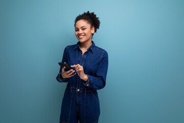 charming slender young hispanic brunette woman with fluffy curly hair in a blue denim suit is chatting in a smartphone