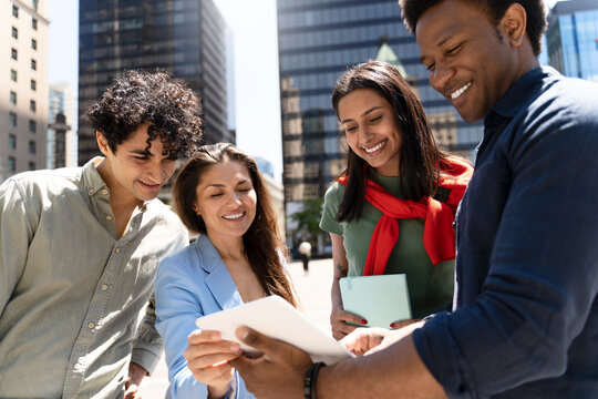 Group of smiling multiracial people using digital tablet, working together on the street. Happy friends watching videos, talking  communication outdoors. Portrait of students, diversity concept 