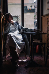 Young girl in white dress drinking coffee in a coffee shop near the window in the sun. Silhouette.