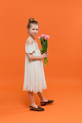 summer dress, preteen girl in white sun dress holding pink tulips on orange background, fashion and style concept, bouquet of flowers, fashionable kid, vibrant colors, summer dress, full length