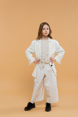 trendy preteen girl in white suit and black shoes looking at camera while standing on beige background, fashionable outfit, formal attire, child model, trendsetter, style, posing with hands on hips
