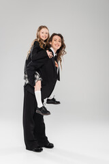 cheerful mother piggybacking her daughter, working mom in business attire and schoolgirl in uniform on grey background in studio, modern parenting, fashionable family, having fun