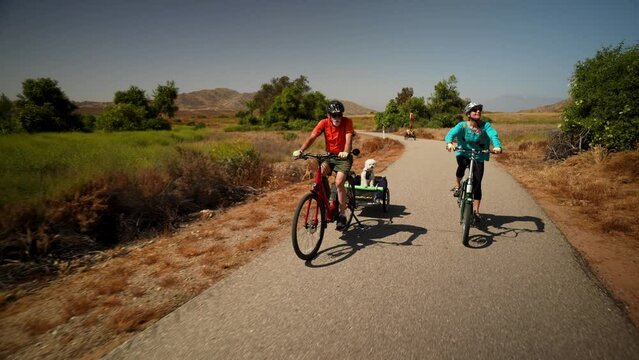Elderly senior man biking on an electric bike on a trail pulling a trailer with a dog in it with a mature woman on an e-bike next to him.