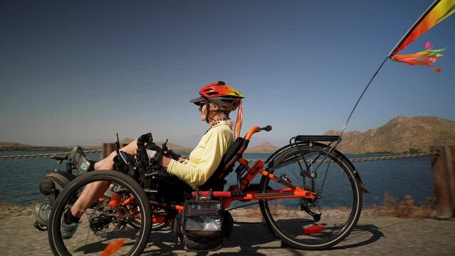 Closeup side view of elderly senior woman riding recumbent e-bike electric tricycle bicycle on a path on a sunny day beside a lake with mountains in the distance.