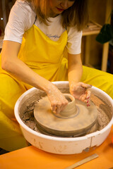 a girl in a yellow apron holds a piece of clay in her hands, sitting in front of a potter's wheel