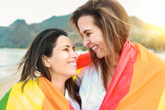 Lesbian gay couple having wearing LGBT rainbow flag with beach on background - Travel summer vacations