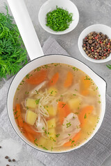 Chicken soup with noodles and vegetables in white pot on a gray concrete background.